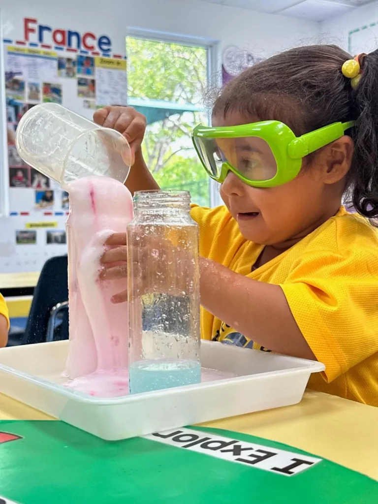 A young student wearing safety goggles conducts a science experiment pouring a bubbling liquid into a bottle